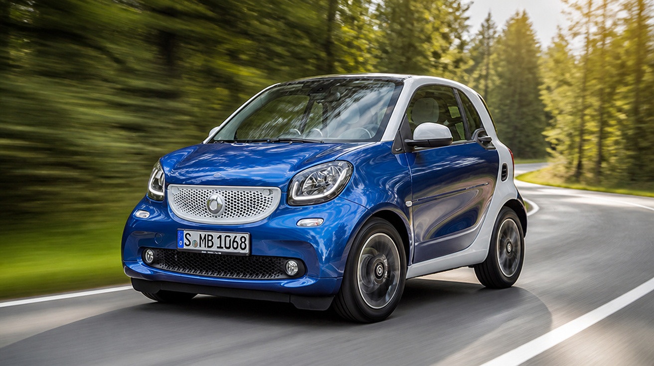 Blauer und weißer Smart Elektro-Kleinwagen fährt auf kurviger Straße in waldreicher Umgebung.