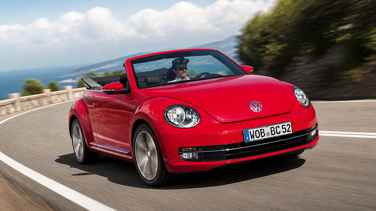 Roter Volkswagen Beetle Cabrio fährt auf kurviger Küstenstraße bei sonnigem Wetter.