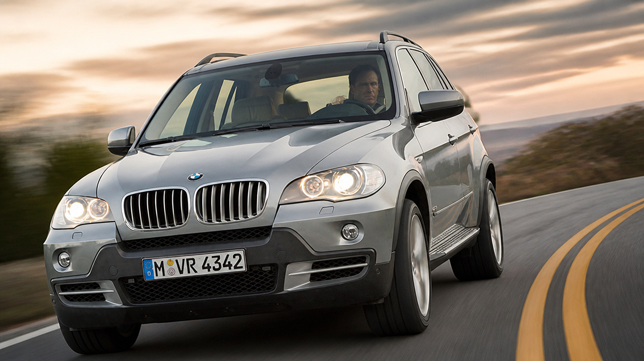 Silberner BMW SUV fährt auf kurviger Straße bei Sonnenuntergang, Fahrer konzentriert am Steuer.