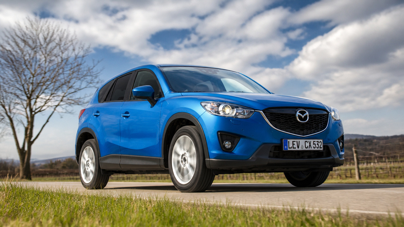 Blauer Mazda SUV fährt auf Landstraße bei bewölktem Himmel und grüner Wiese, Baum im Hintergrund.