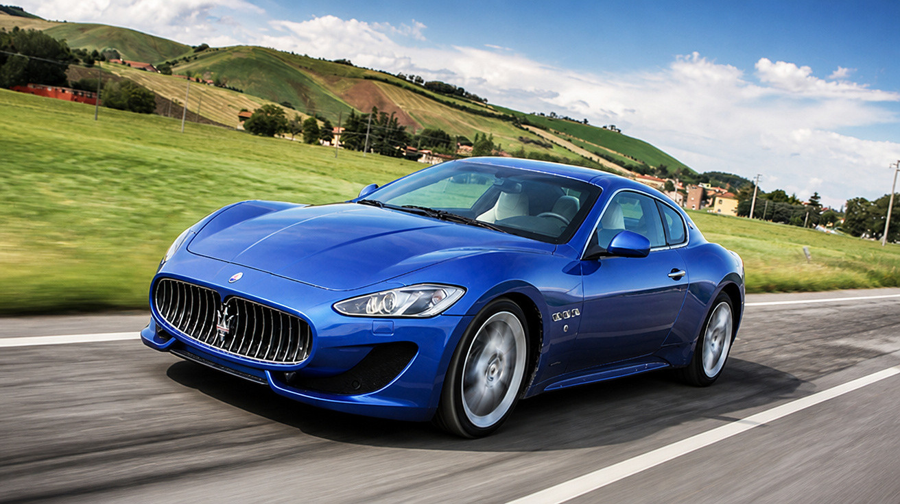 Blauer Maserati Sportwagen fährt auf Landstraße mit grünen Hügeln und blauem Himmel im Hintergrund.