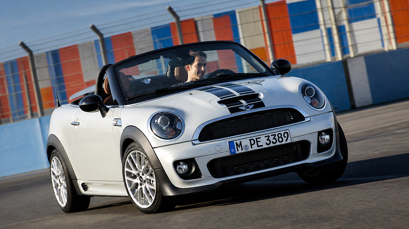 Weißer Mini Cabrio mit schwarzen Streifen fährt auf Rennstrecke, Fahrer lächelt konzentriert