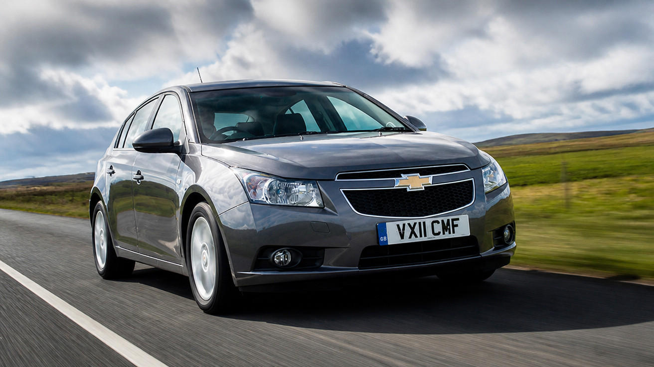 Grauer Chevrolet Limousine fährt auf einer Landstraße bei bewölktem Himmel und grünen Feldern.