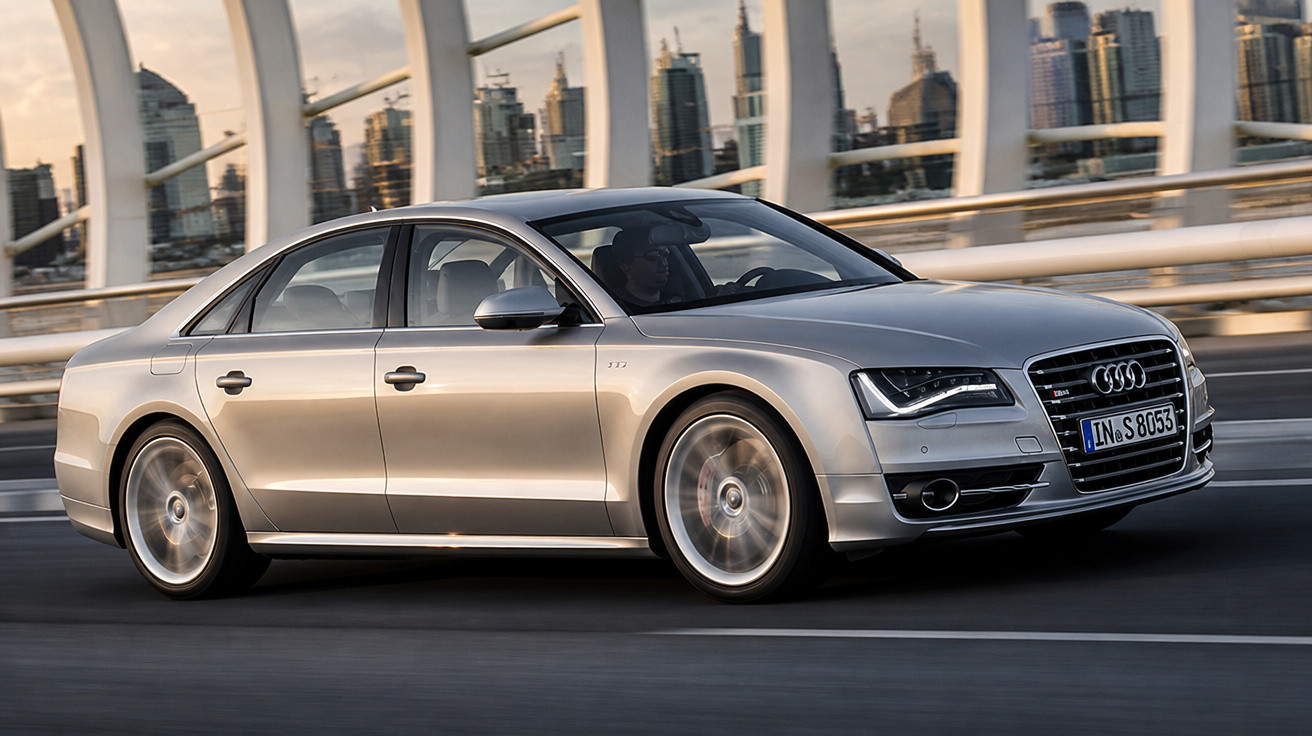 Silberner Audi Limousine fährt bei Sonnenuntergang auf urbaner Brücke mit moderner Skyline im Hintergrund.