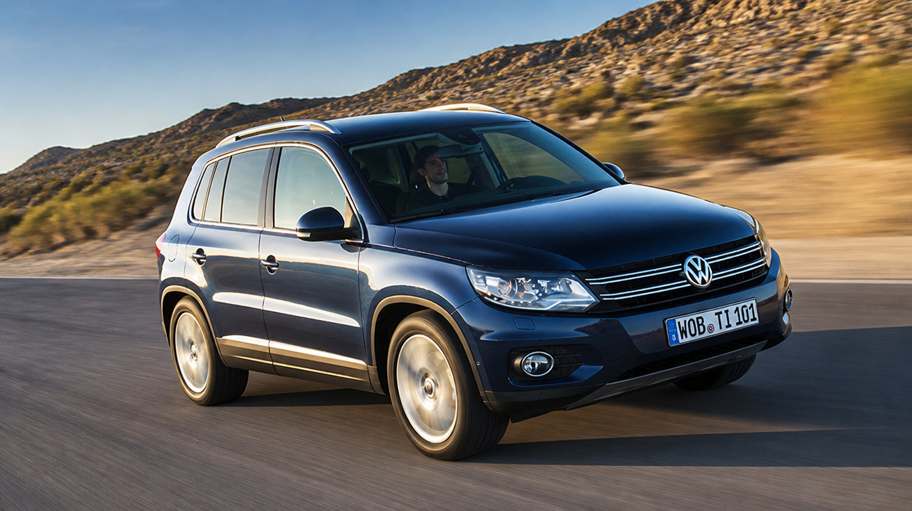 Blauer Volkswagen SUV fährt auf einer Straße mit bergiger Landschaft im Hintergrund bei Sonnenschein.