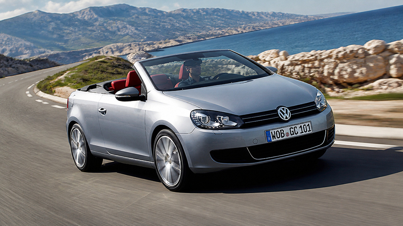 Silbernes Volkswagen Cabrio fährt auf kurviger Küstenstraße mit Berg- und Meerblick im Hintergrund.