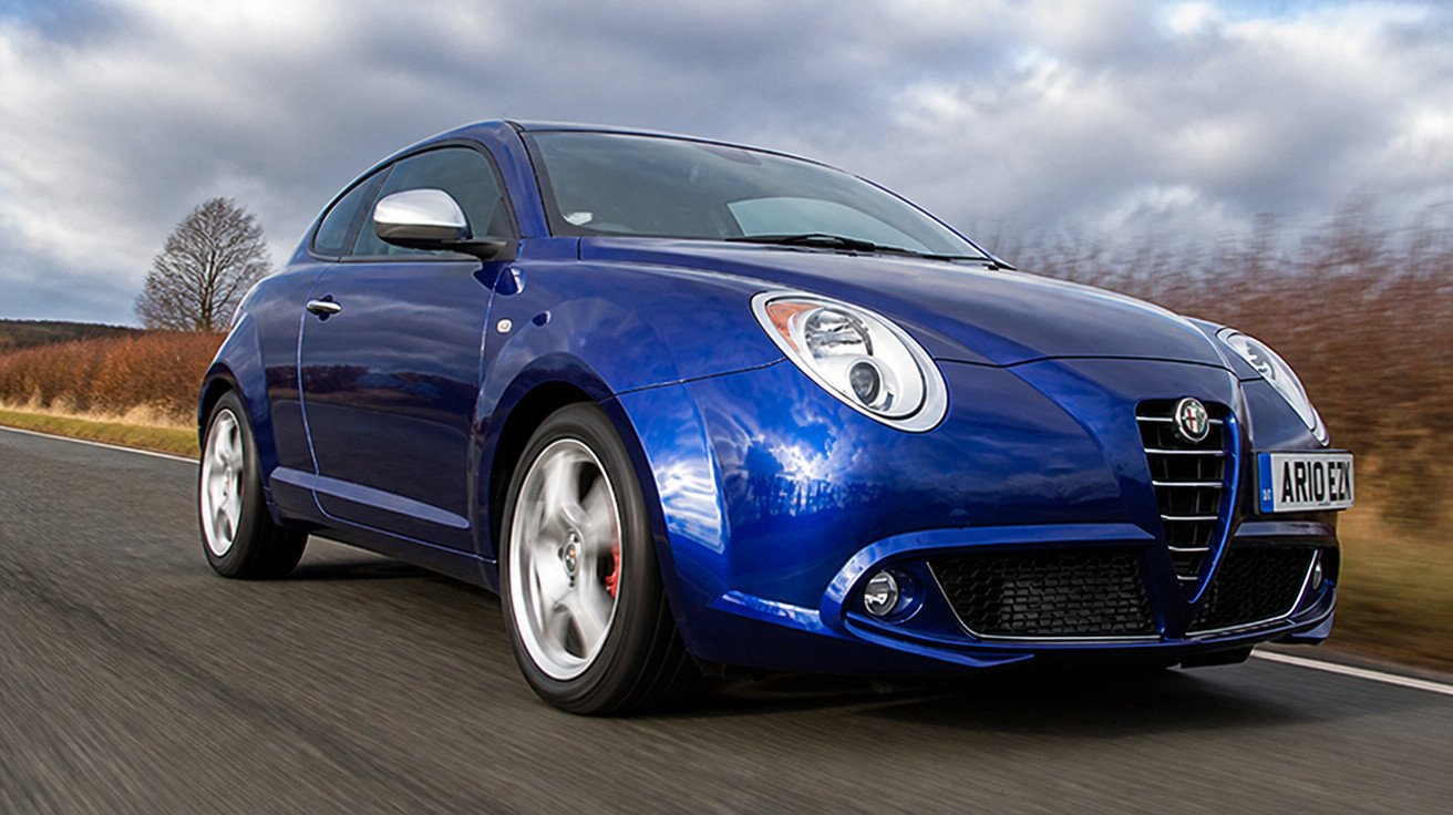 Blauer Alfa Romeo Kleinwagen fährt auf Landstraße unter bewölktem Himmel.