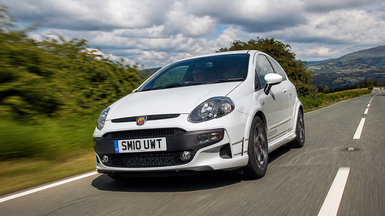 Weißer Abarth Kleinwagen fährt auf einer Landstraße bei bewölktem Himmel in ländlicher Umgebung.