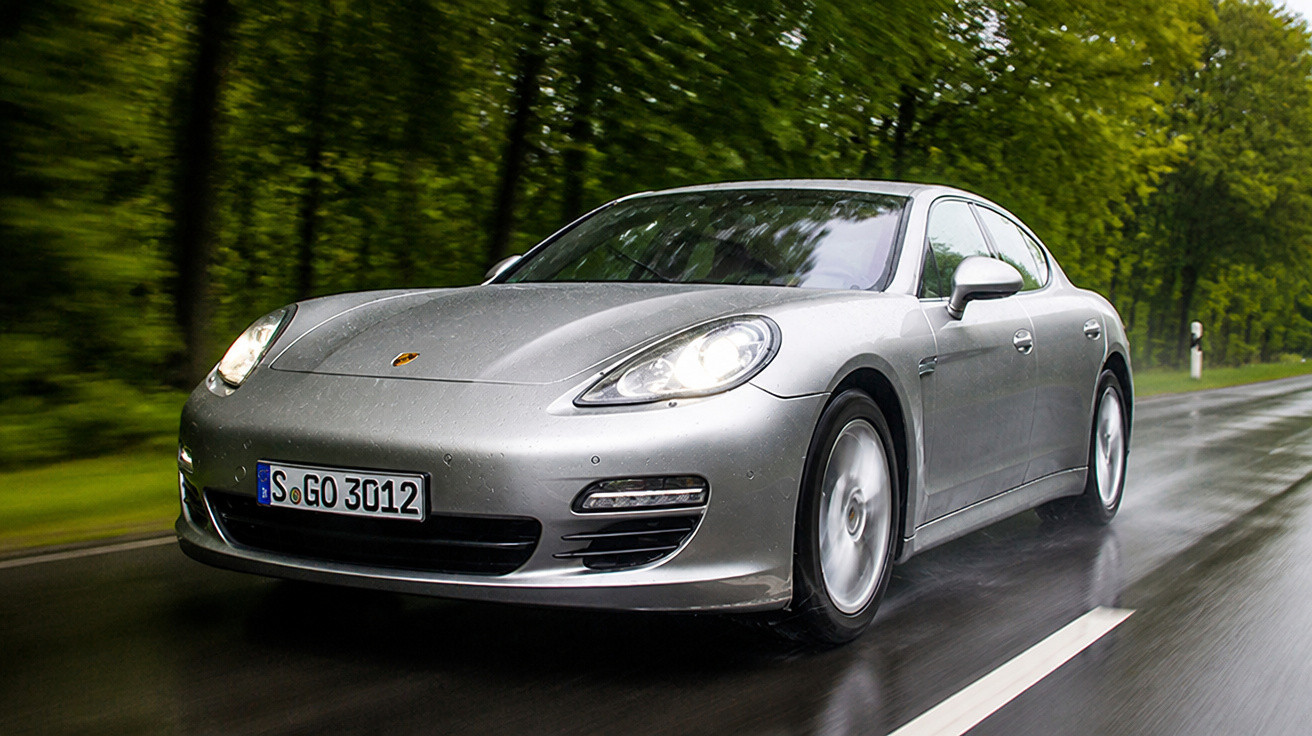 Silberner Porsche Panamera fährt bei Regen auf nasser Landstraße mit grünen Bäumen im Hintergrund.