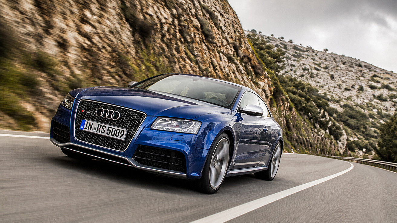 Blauer Audi RS 5 fährt auf kurviger Bergstraße mit Felswand und bewölktem Himmel im Hintergrund.