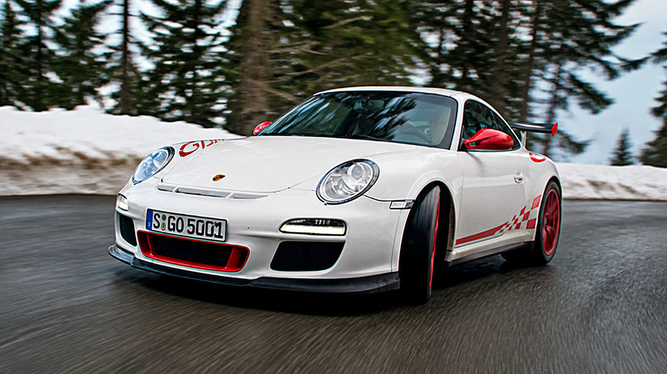 Weißer Porsche 911 mit roten Akzenten fährt auf kurviger, nasser Bergstraße mit Schneeresten am Straßenrand.