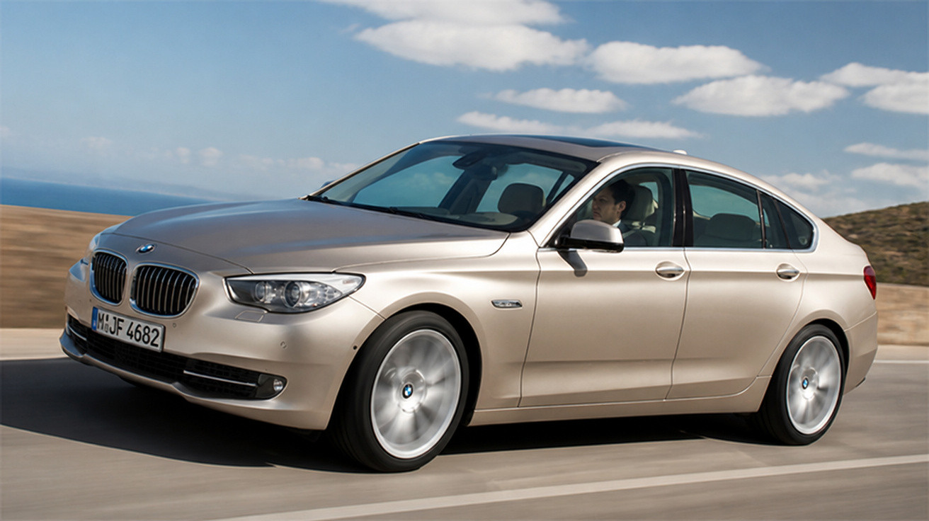 Beige BMW Limousine fährt auf Straße bei klarem Himmel, Fahrer sichtbar am Steuer.
