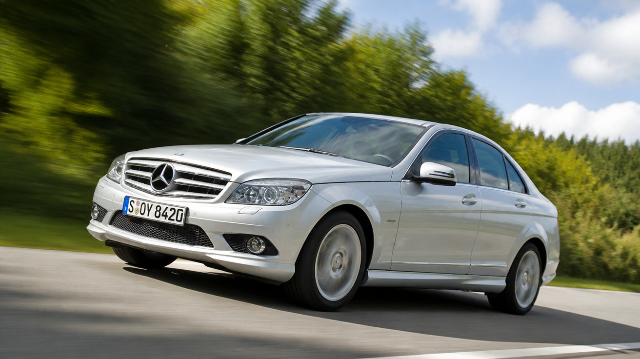 Silberner Mercedes-Benz fährt auf einer Landstraße mit grünen Bäumen im Hintergrund bei sonnigem Wetter.