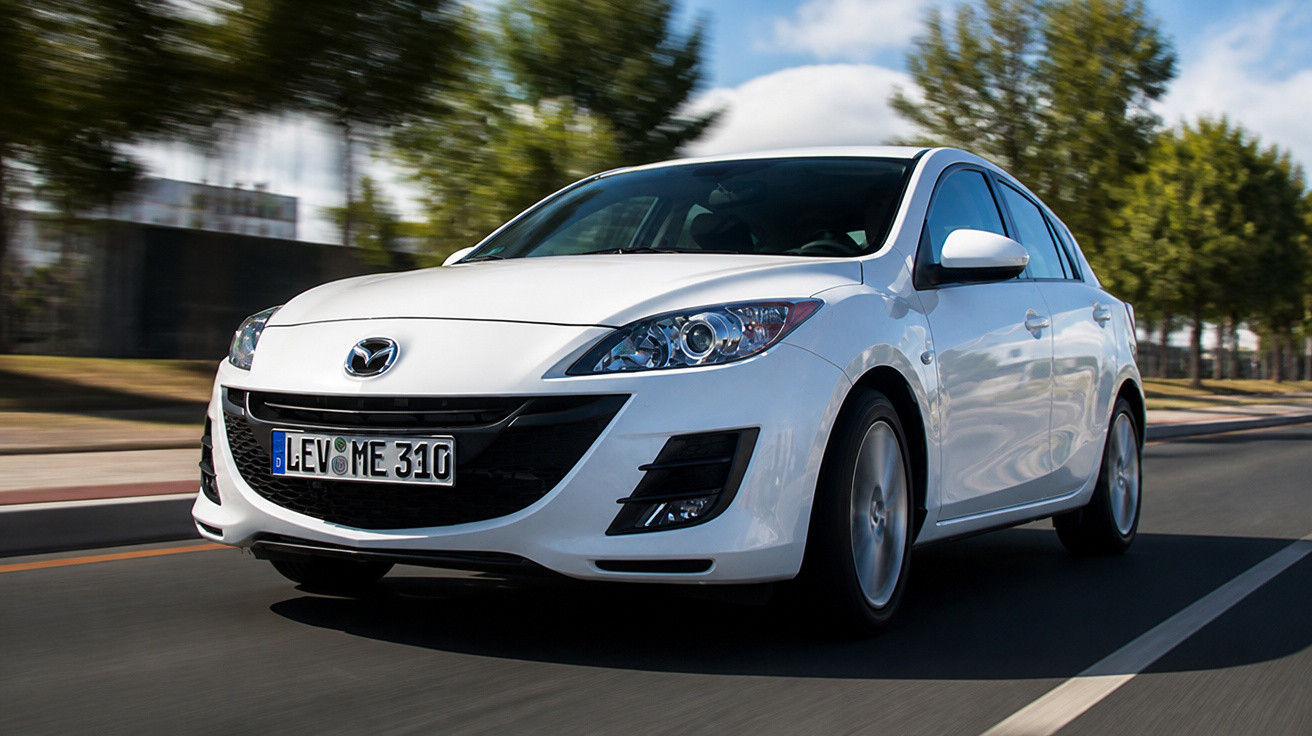 Weißer Mazda 3 fährt auf einer sonnigen Straße mit Bäumen im Hintergrund.