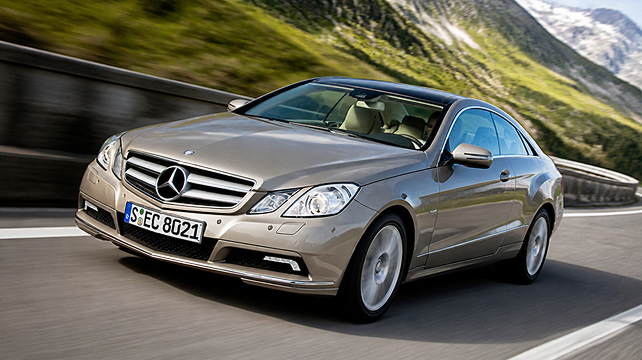 Silberner Mercedes Coupé fährt auf kurviger Bergstraße bei sonnigem Wetter.
