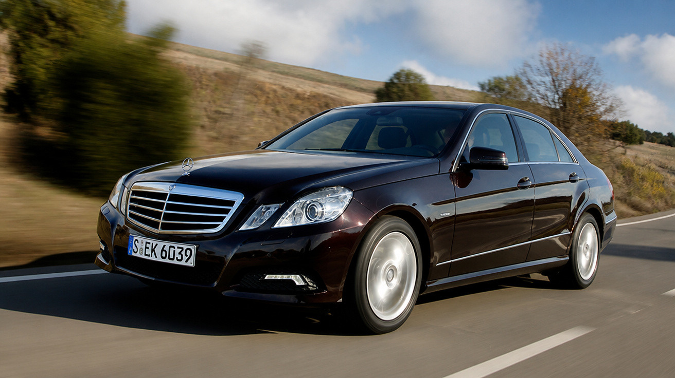 Schwarze Mercedes-Benz Limousine fährt auf Landstraße bei blauem Himmel und leichter Bewölkung.