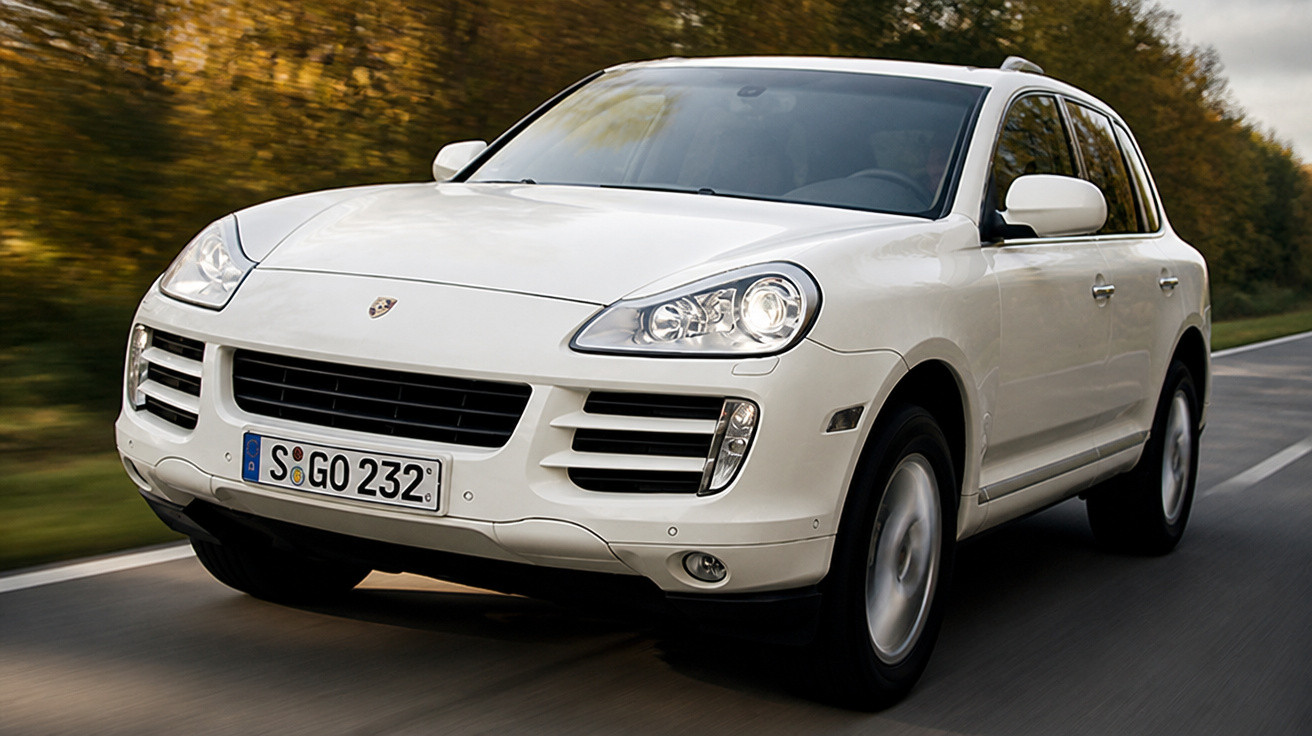 Weißer Porsche SUV fährt auf Landstraße, herbstliche Bäume im Hintergrund, Nahaufnahme Frontansicht.