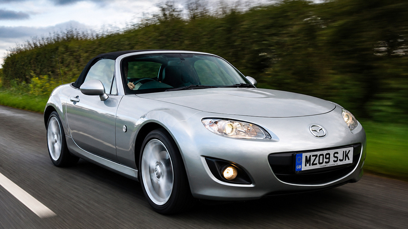Silberner Mazda MX-5 Roadster fährt auf Landstraße bei bewölktem Himmel.