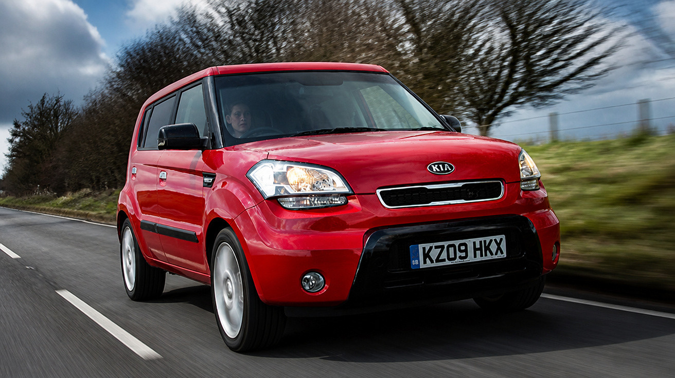 Roter Kia Soul fährt auf Landstraße bei bewölktem Himmel, Bäume im Hintergrund.