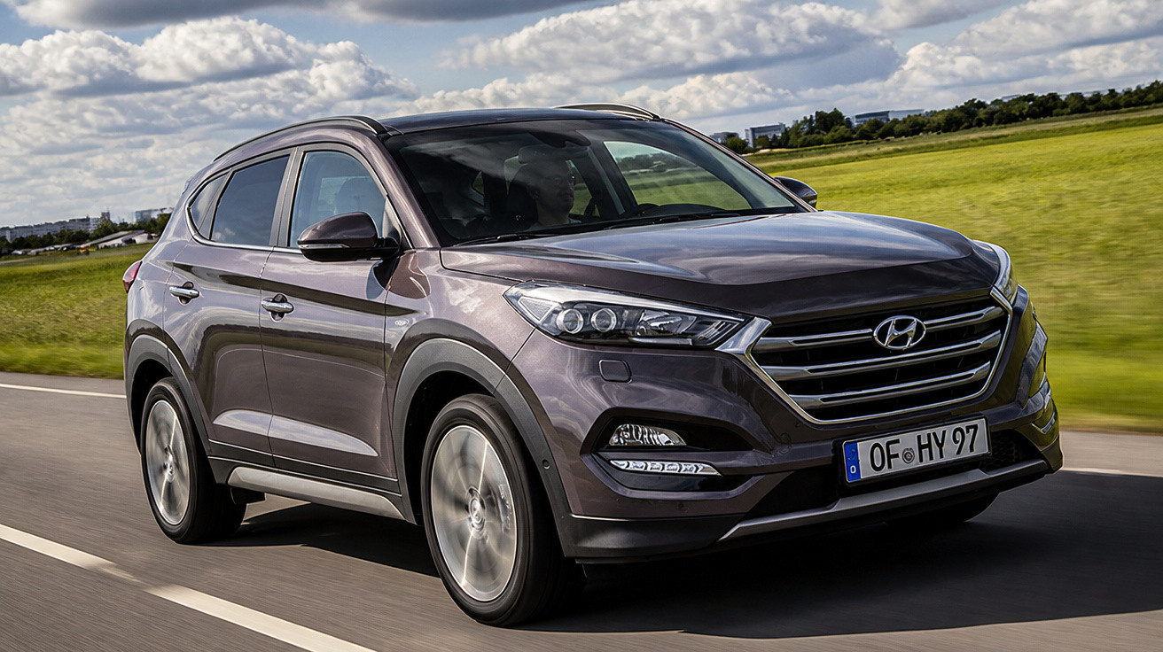 Hyundai SUV in Dunkelgrau fährt auf Landstraße bei bewölktem Himmel, grüne Wiesen im Hintergrund.