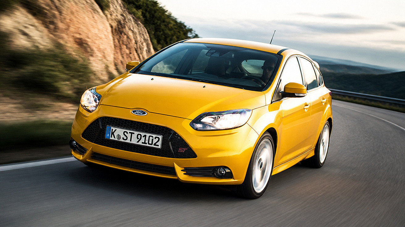Gelber Ford Focus ST fährt auf Bergstraße mit Felsen und bewölktem Himmel im Hintergrund.