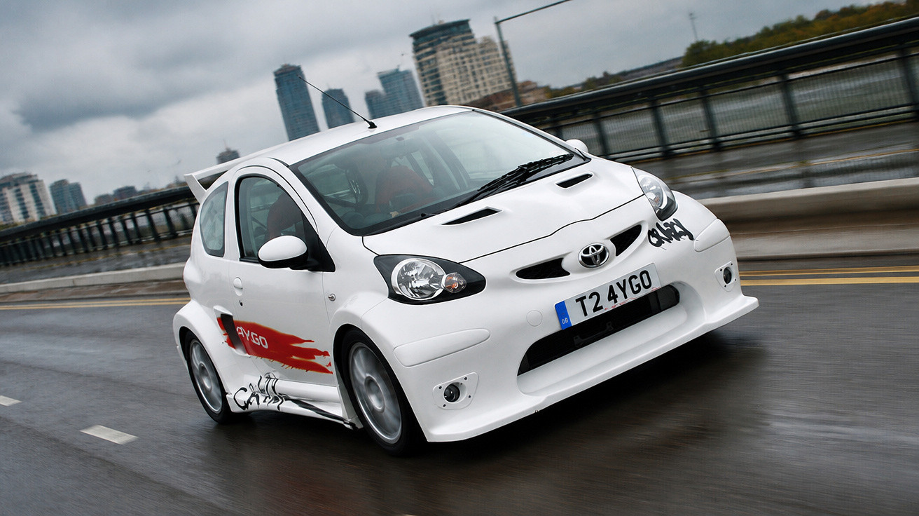 Weißer Toyota Aygo fährt bei nassem Wetter auf einer Brücke in der Stadt.