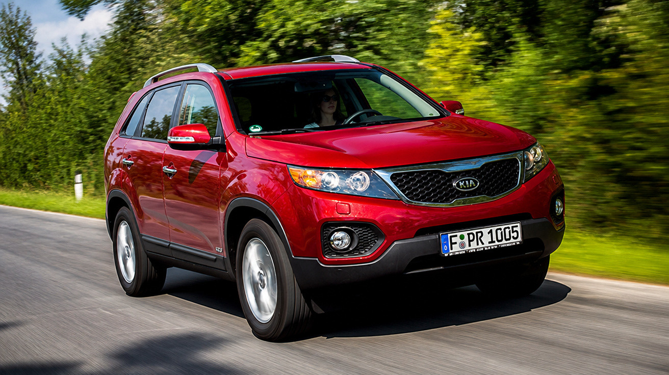 Roter Kia SUV fährt auf einer Landstraße, umgeben von grünem Laub und Bäumen bei Sonnenschein.